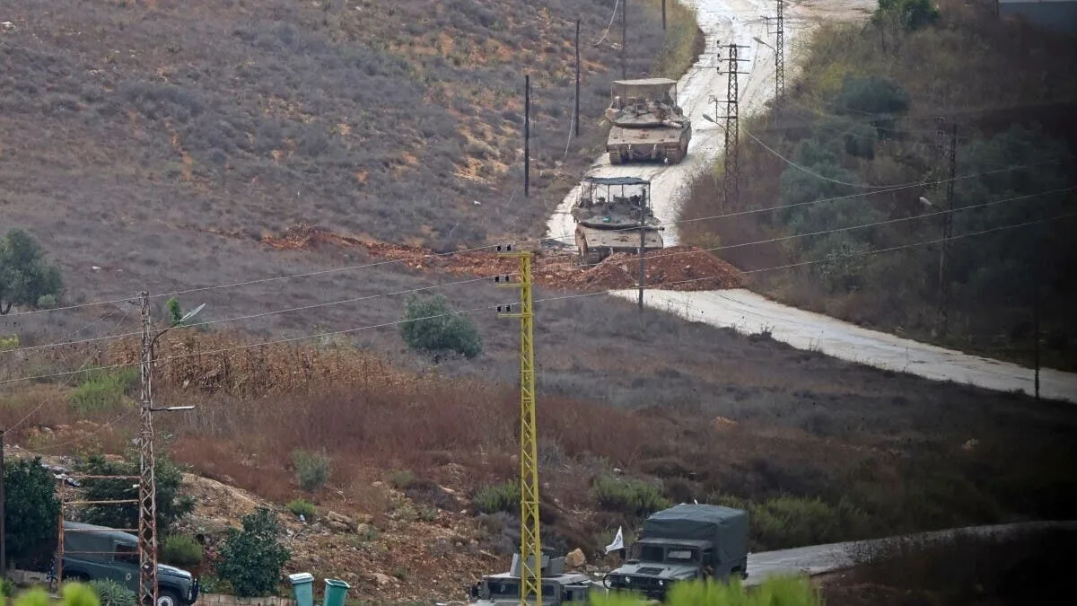 Military armored vehicles travel along a rural road in the Deir Mimas area, amid dry terrain and power poles.