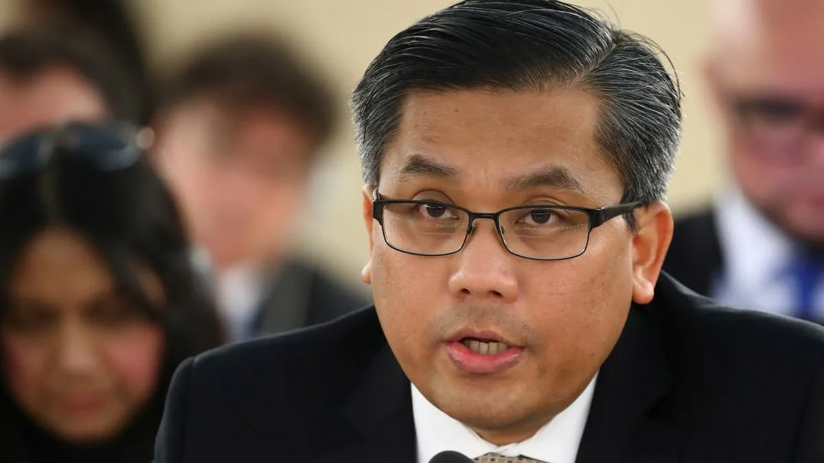 Close-up of a man wearing a suit and glasses speaking during a meeting, with other people visible in the background.