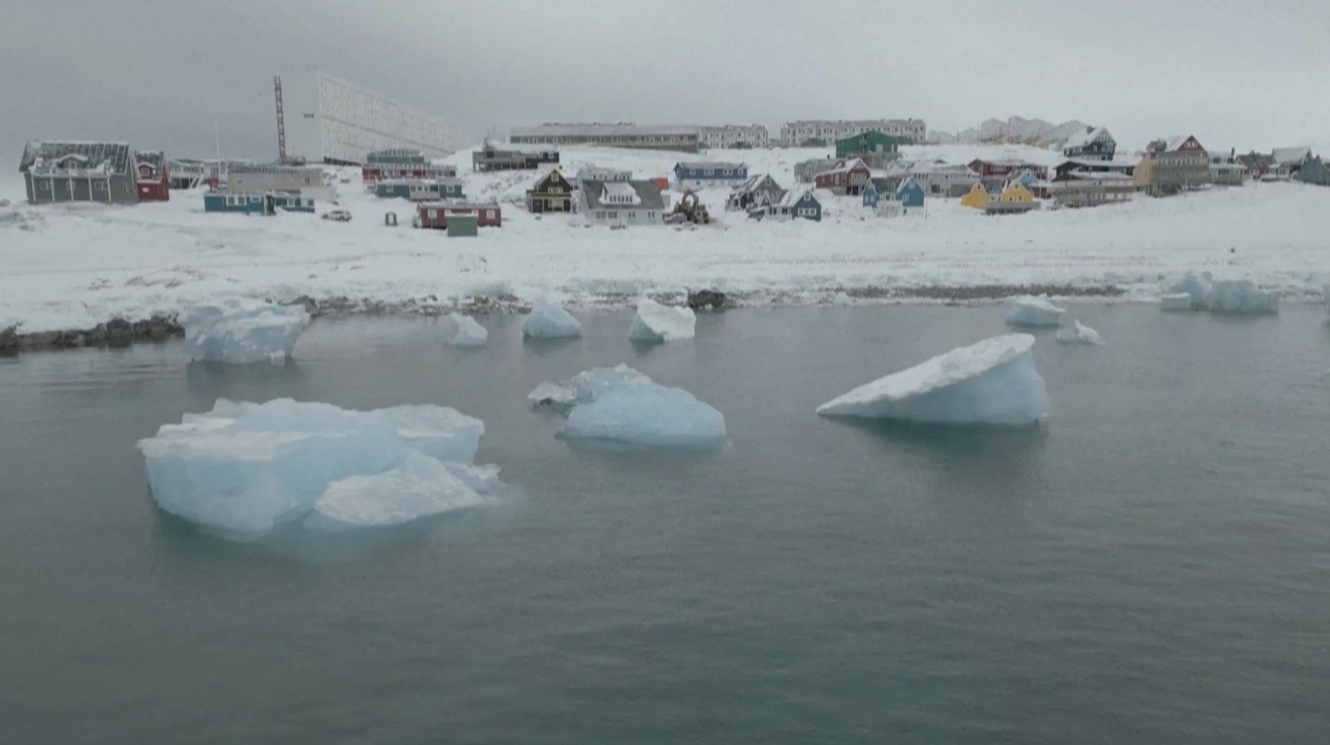 A snow-covered coastal settlement in Greenland, with floating pieces of ice in the water in the foreground.