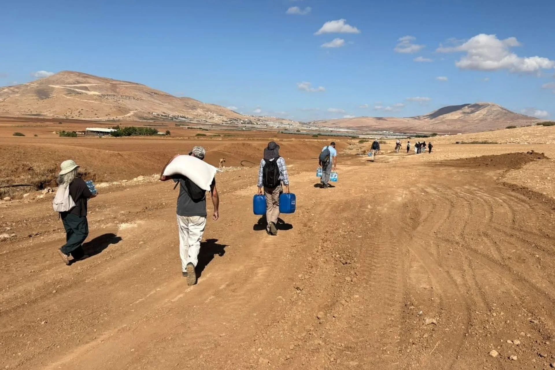 People walk along a dirt track in an arid area carrying water containers and belongings. Fields and barren hills are visible in the background at Ras al-Ahmar in the northern Jordan Valley.